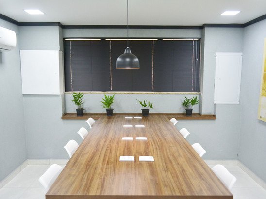 Sala de Reunião White com mesa longa de madeira, cadeiras brancas e plantas junto à janela